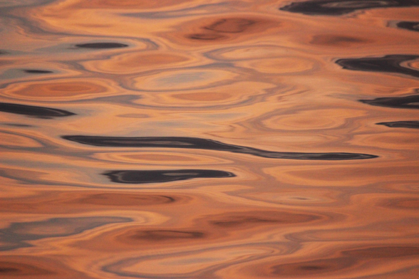 Photograph of sunset light reflected on a lake's surface forming ovals and swirls of tan, brown, lavender, and grey.