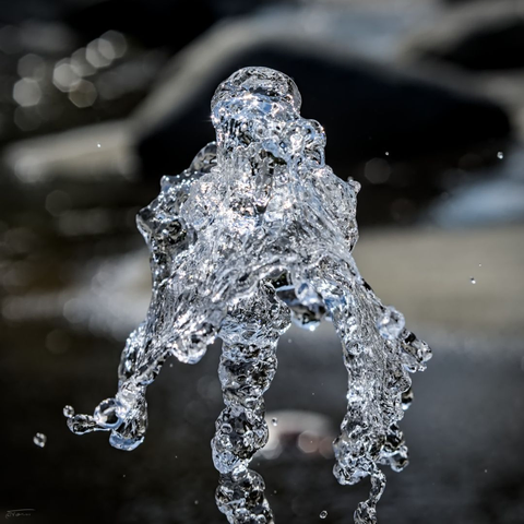 Das Bild zeigt einen Moment, in dem Wasser in einer ungewöhnlichen, fast skulpturalen Form eingefangen wurde. Es sieht aus, als würde das Wasser kurz "stehenbleiben" und eine abstrakte, fast figurative Gestalt bilden — fast wie eine kleine, fließende Silhouette. Die Bewegung des Wassers ist so, dass es in dieser Aufnahme wie eine glitzernde, transparente Skulptur wirkt. Der Hintergrund ist unscharf, was die dynamische Form des Wassers noch stärker betont.
