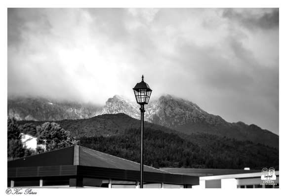 A moody black and white photograph featuring a classic street lamp centered against a dramatic mountain landscape, in Queenstown.

The foreground shows the roofline of a modern building. Behind the light, a forested slope rises to meet rugged, rocky peaks shrouded in low-hanging, textured clouds.

The overall composition creates a strong contrast between the man-made structure and the raw natural environment.