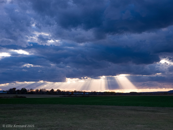 A scene across a dark field showing the sun's rays coming down through dark clouds.