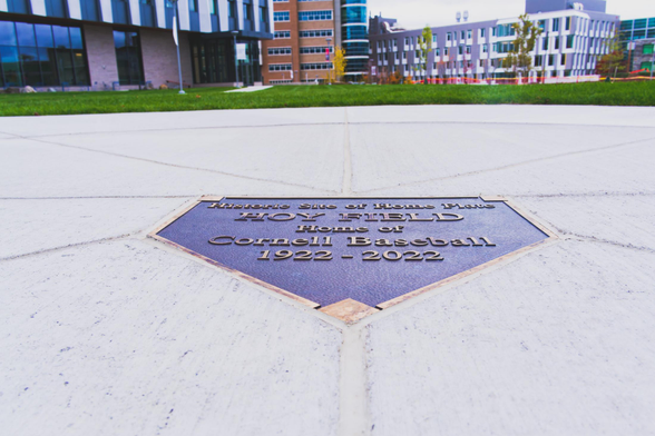 A plate in the shape of a home plate reads: Historic Site of Home Plate HOY FIELD Home of Cornell Baseball 1922-2022 in the middle of a round cement circle with grass beyond and several buildings further away