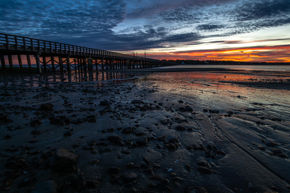 A photograph sunset over the long, wooden Powder Point Bridge. The tide is low and the sun is reflecting off the mud and sand in the foreground.
