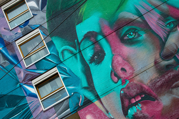 The side facade of an apartment building, photographed diagonally from below. On the left, three square windows, one below the other. A mural covers the entire facade, primarily in blue, sea green, and purple. Across the entire right side, next to the windows, is a woman's face, also in purple, blue, and green. Her mouth is slightly open, teeth visible, and the corners of her mouth are turned down. It looks dramatic. It strikes me as a bit creepy.