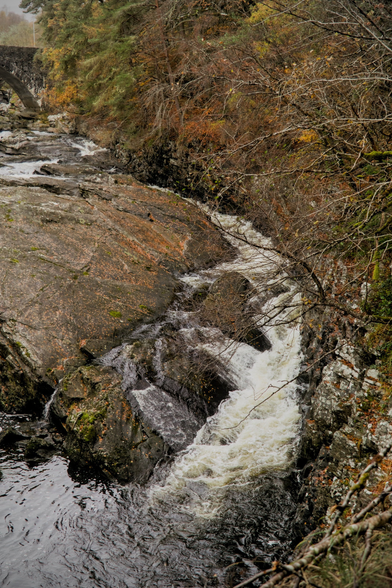 Portrait format photo of a part of a river making eddies in a smooth curve right around a big rock in the middle. The colours are autumnal with orange trees in the top right. A piece of a stone bridge can be seen in the background top left