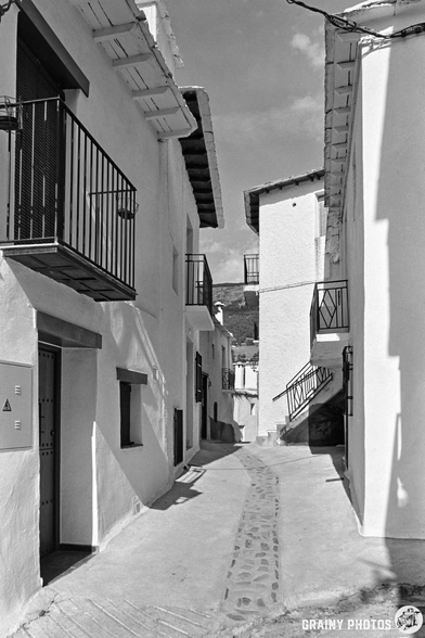 A narrow cobbled street in Capileira, lined with whitewashed buildings featuring balconies, stretches under a clear sky; mountains can be seen in the distance.