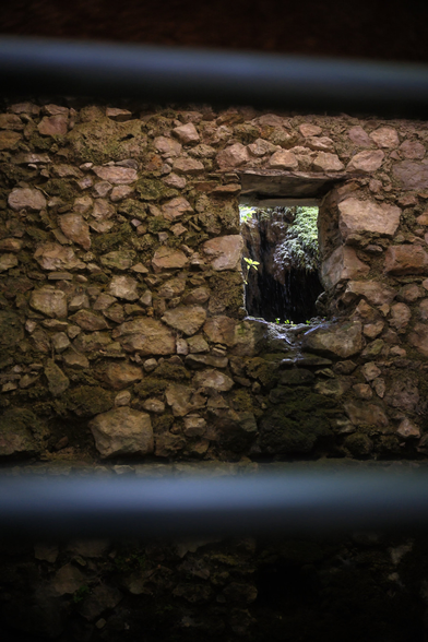 Portrait format photo with in the foreground, 2 horizontal metal bars, out of focus, framing a wall made of reddish stones with a rectangle hole in it, an old window maybe, through which we can see green vegetation