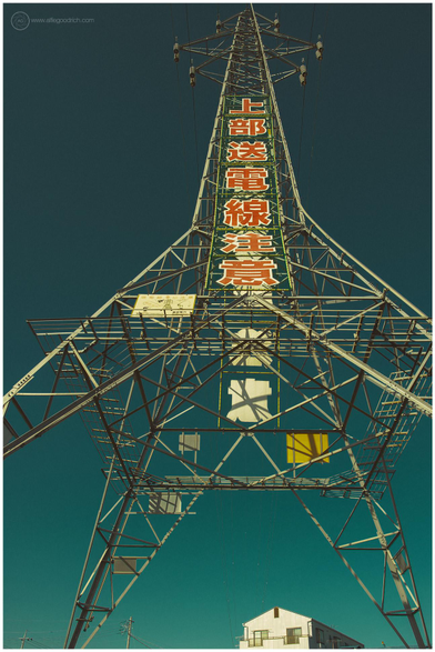 Electricity pylon with a house underneath it in the background. Deep blue, cloudless sky.

Text in Japanese on the pylon says "Beware of overhead electricity cables".