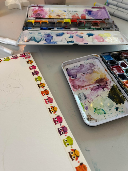 Foto: Auf meinem Kunst Tisch stehen die aufgeklappten Farbkästen mit Aquarellfarben und ein an den Rändern mit Washi Tape abgeklebtes weißes Blatt Papier liegt daneben. 