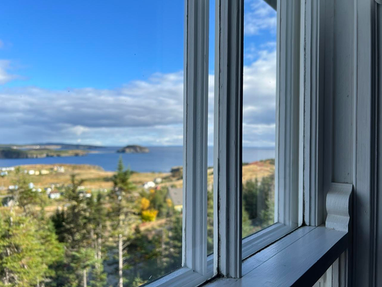 Blurred coastal ocean landscape viewed at an angle through a paned window with a painted white wooden frame. The glossy paint reflects indirect afternoon sun, and with the contours and ridges of the wood emphasized from shadows. The coastal scene shows trees and houses, distant but in the foreground, with rugged coast and island in the background under a bright blue sky with a bank of dense white clouds.  [Port Rexton (NL, Canada), October 2025]