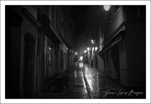 This image captures a narrow, cobblestone street at night, illuminated by a series of warm street lamps that create pools of light on the wet surface below. The wetness suggests recent rain, giving the pavement a reflective quality that enhances the moody atmosphere. The street is flanked by old buildings with shuttered windows and small balconies, evoking a historic European town or village.
The solitary figure walking at the center, slightly blurred due to motion or rain, adds a sense of loneliness or introspection, contributing to the image’s contemplative mood. The black and white treatment strips away color distractions, focusing our attention on texture, light, and shadow, emphasizing the timelessness and quiet solitude of the scene.
Symbolically, the image could be interpreted as a journey through an uncertain or solitary path, with the lights symbolizing hope or guidance amid darkness. The narrow, enclosed street alludes to constraints or a focused direction, while the reflection of light on water adds a layer of ephemeral beauty.
Artistically, the image reflects themes common in noir photography or cinema, where light and shadow interplay to evoke mystery or melancholy. The artist’s signature at the bottom right anchors the image as a crafted work, highlighting its intention as a piece of visual storytelling rather than a mere snapshot.
