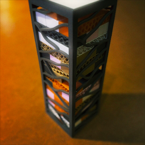 A 3D-printed tabletop Jenga tower stands with its corrugated pieces' internals exposed under a small, tungsten interior light, all in hues of brown, orange, gray, and white.