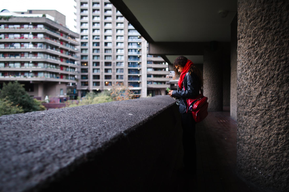 The image captures a moment in an urban environment, likely a residential area. The primary subject is a person, a woman, standing on a balcony or a raised walkway, gazing at a mobile phone in her hands. She is wearing a black leather jacket, a vibrant red scarf that contrasts with her dark hair, and black pants. A bright red bag hangs on her shoulder.

The architectural setting is dominated by Brutalist concrete buildings. In the background, there are several large apartment buildings with many windows and balconies. The buildings have a uniform, rectangular design, typical of Brutalist architecture. The color palette of the buildings is primarily shades of grey and beige, with some red accents visible on the balconies and windows.