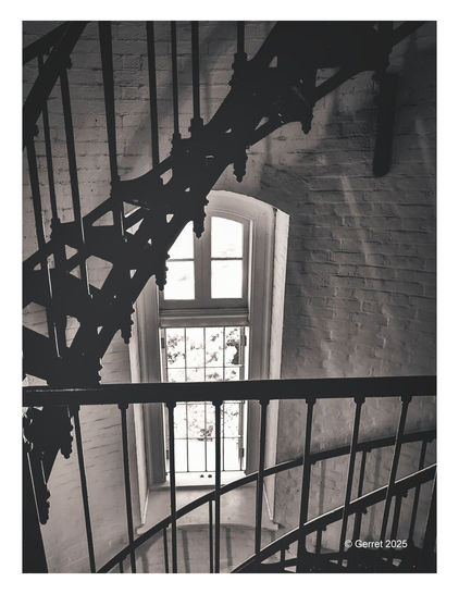 Black and white image of a spiral staircase winding around a tall window. Light streams through, casting shadows and creating a mysterious, historic ambiance.