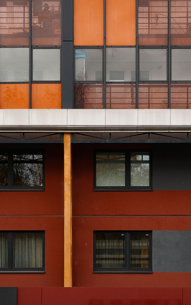 The photo shows a part of a modern building with large balcony windows and a red and orange wall. Ordinary windows with black frames reflect the surrounding environment.