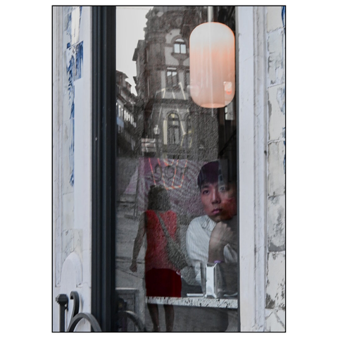 The image shows a view from outside through a glass window into a cafe on an urban street. Inside the window, a person is partially visible, sitting behind the glass and looking outward. The person is wearing a white shirt and a cap.

The reflection and view of the street are visible in the window. A person in a red dress is walking on the sidewalk. The street features buildings with architectural details, including arched windows and stone facades. The scene is captured during daylight, and the glass surface of the window creates a layered effect with reflections and transparency. (draft by Mistral.ai, edited by author)