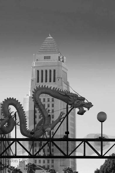 This is a black and white cityscape photo in portrait format of the Los Angeles Chinatown Gateway and the LA City Hall (2014).
In the foreground, near the base of the image is a lattice overhead gantry spanning a city street. Mounted on top of the gantry is an large, long, ornate sculpture or model of a serpentine Dragon that is curled over the gantry. The tail is to the left, the large and fearsome head facing to the right and positioned to the right of centre. Our mythical creature has a narrow, scaly body with sharp,  triangular dorsal spines in the back. The raised head is large with bulging eyes, and an open mouth with large teeth. In the background is the iconic Los Angeles City Hall. 140m (450ft) tall with 32 floors and has been destroyed on film by more invading extraterrestrials and assorted monsters than appear in a whole series of Star Trek. 