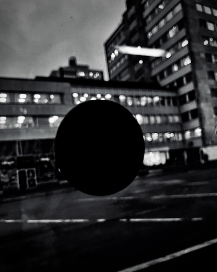 A blurred black circle in the foreground with illuminated buildings behind, captured in moody black and white at dusk.