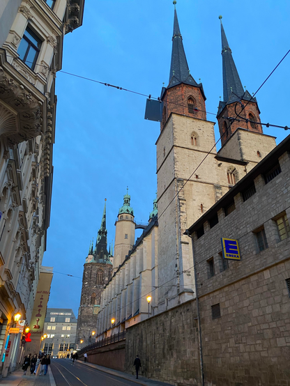 A scene in a city centre.
On the right side of the photo there is a big church made of sandstone with two towers top right (made of red bricks) and two towers in the centre (of the picture).
Behind the church there‘s a gothic tower in the distance (lower centre left).
On the left side there‘s a row of house next to a street and opposite of the church.
On the street there are tram tracks.
A couple of people are walking on the pavement in the distance (lower left and centre).
The street leads to an illuminated department store.
Above the buildings there‘s a blueish cloudy autumn sky.