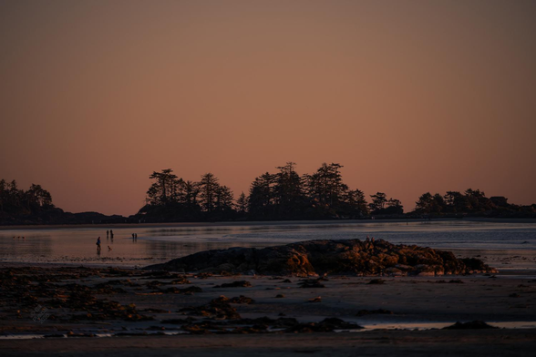 Looking out toward some islands just offshore - silhouetted rock and trees. A few people are wandering on the near shore, watching the sunset. The colours are low and saturated dusky pinks and oranges fading into purple and blue.

Traditional territory of ƛaʔuukʷiʔatḥ (Tla-o-qui-aht) First Nations people. 