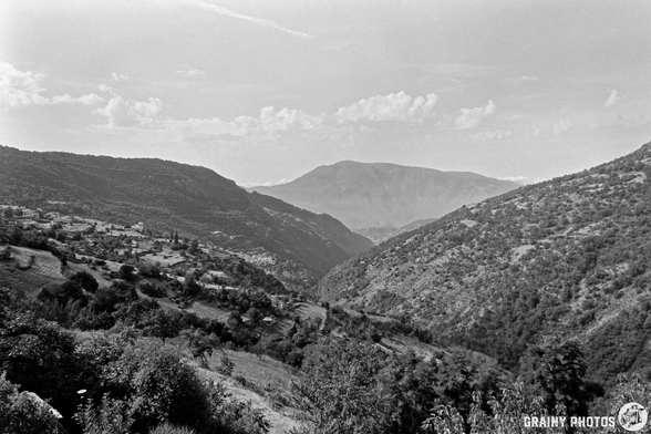 Black and white photo of the Poqueira Valley’s mountainous landscape with rolling hills and a distant peak under a partly cloudy sky. Vegetation and scattered trees cover the slopes.