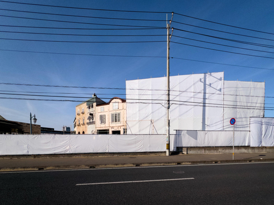 An empty road in the foreground, blue sky beyond. A single post carrying twelve or so power lines cuts the scene two thirds of the way from the left edge. Behind it a building which is partially shrouded in white awnings in readiness for demolition. The front of the building which is still visible comprises a two story curving facade in pastel shades. It was a shop made to look like an old town house. There’s a hint of a Disney arcade about the architecture. The structure and its many windows are now no more.