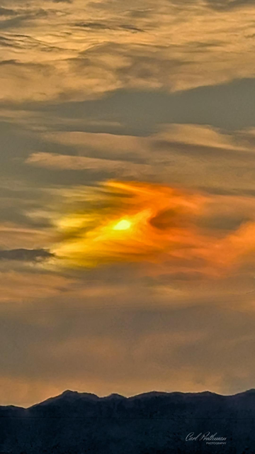 A close-up view of glowing orange and yellow clouds during sunset above a dark mountain ridge. The sunlight forms a fiery swirl within the cloud layers, blending warm tones into the muted blue-gray sky. The overall feeling of the photo is calm yet powerful, like nature showing a brief moment of quiet intensity.
