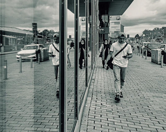 Ein junger Mann in Jeans und T-Shirt hält den Blick fest geradeaus und sein Handy in der Hand, während er an einem verglasten Gebäude auf den Fotografen zu schreitet. Er spiegelt sich zusammen mit anderen Fußgängern und der gegenüberliegenden Straßenseite im Glas der Fensterfront. 
.
A young man in jeans and a shirt keeps his gaze firmly ahead and his mobile phone in his hand as he walks towards the photographer along a glass-fronted building. He is reflected in the glass window front along with other pedestrians and the opposite side of the street. 
.