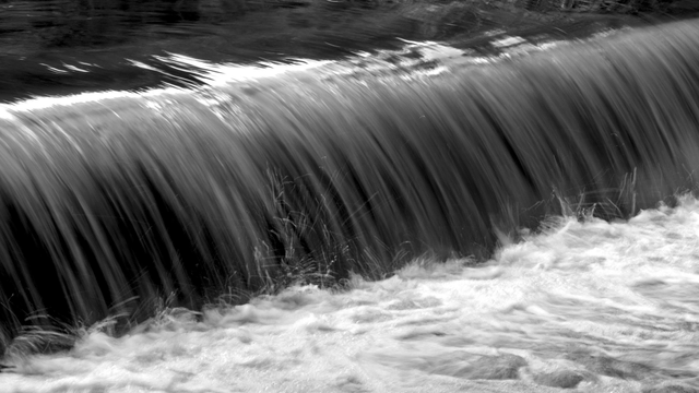 A black and white diagonal shot of a small waterfall, with its front going from the lower left to the upper right side of the image. The water is drawing lines due to a slightly longer exposure, and there are tiny drops in the foreground, drawing white streaks.
