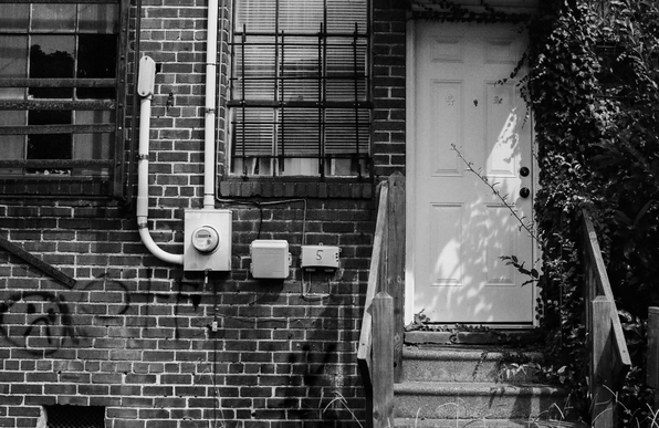 A black and white photo of the back of a brick building showing two iron barred windows, one with some glass panes broken out.  There's also a white wooden door and concrete steps leading to ground level.  Electric boxes and graffiti are on the brick wall and climbing vines on the right side of the door.