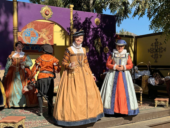 Two ladies in perfect Renaissance period costumes smile for the camera. Others are in the background.
