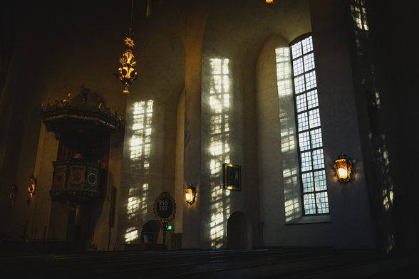 A photograph from the inside of a tall church. The photo is taken from the central aisle towards one of the side walls. To the right is a tall vertical window, casting light on the wall of the niche it sits in , showing shadows from the window grid and from a tree outside.

Next to it to the left is another window in a niche, but the windows itself is obscured, only the light on the niche wall from it is visible. Next on the same wall is a raised decorated pulpit. on the wall are also some lamps and from the ceiling another lamp hangs down.

Inside the church is quite dark, clearly highlighting the incoming light from the windows.