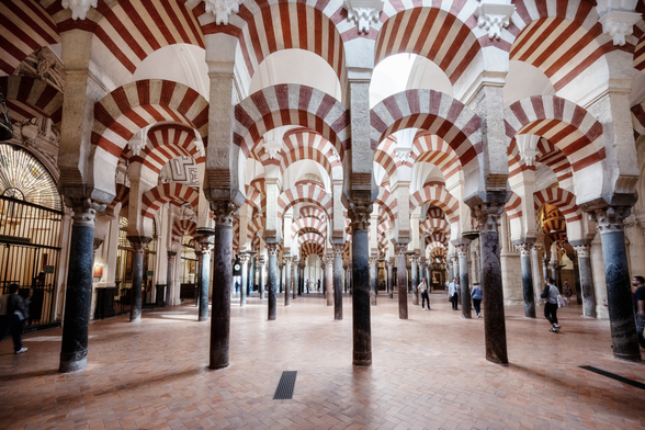 Der Innenraum der Moschee-Kathedrale von Córdoba. Eine unendliche Reihe dunkler Säulen trägt die berühmten rot-weissen abwechselnd gestreiften Hufeisenbögen, die sich in die Tiefe des Raumes erstrecken. Der Boden ist hellbraun gefliest. Wenige Besucher beleben die riesige Halle.