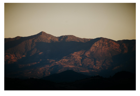 Golden light bathes a rugged mountain range during sunrise or sunset, casting dramatic shadows across ridges and valleys. The foreground hills appear dark, while distant peaks glow with warm tones, emphasizing the terrain’s depth and texture.