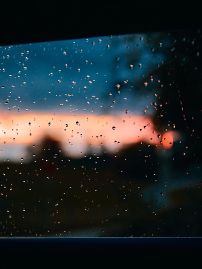 A car window with rain drops on it and a blurred sunset in the background.