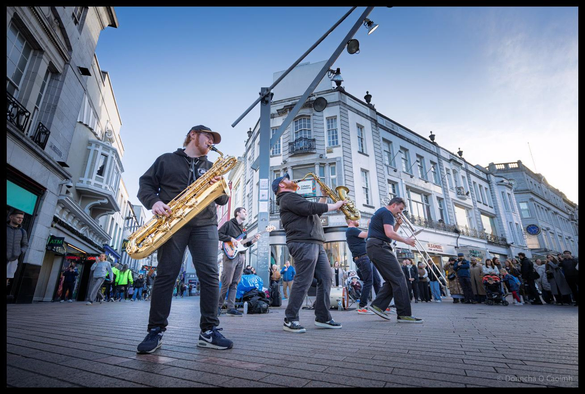 Wide-angle shot of TBL8 Brass Band performing on St Patrick Street during Cork Jazz Festival, with three horn players in foreground including saxophonists and trombonist, Georgian buildings and gathered crowd visible in background under evening sky.
