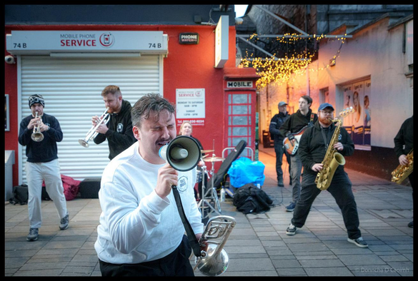 TBL8 Brass band performing on Oliver Plunkett Street in Cork during the Jazz Festival, with frontman on a megaphone in white clothing, surrounded by brass section and horn players in black, with red mobile phone shop and Christmas lights visible in the background at dusk.