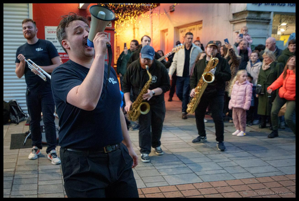 TBL8 Brass frontman performing passionately with megaphone on Oliver Plunkett Street during Cork Jazz Festival at dusk, with band members and large crowd of spectators visible behind him, including families and children, with illuminated shop windows in the background.