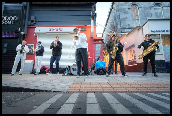 Wide-angle low perspective shot of TBL8 Brass band performing on Oliver Plunkett Street during Cork Jazz Festival, showing the full brass section spread across the pedestrianised street with Georgian buildings, shop fronts, and Christmas lights in the background at dusk.