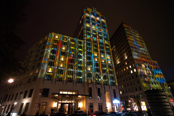 Das Ritz-Carlton in Berlin und sein Nachbarhaus von unten fotografiert. Zwei Gebäude mit je 18 Etagen, deren Fassaden mit einem bunten Motiv angestrahlt werden, vor denen Autos stehen und Leute laufen. Einige Lichter in den zahlreichen Fenstern versuchen, gegen die Muster anzuleuchten.