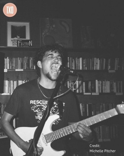 A tall bookshelf labeled "Fiction" stands behind a young masculine-presenting person singing at the top of their lungs into a microphone as they play an electric guitar.