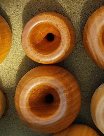 Top-down view of two mouthpieces for alphorns, which lie close together and form a figure eight. Their shape is circular with a flattened surface. It has a recess in the middle that opens into a hole. The mouthpieces are hand made of reddish wood with a beautiful horizontal grain. To the right and left of the two objects, additional mouthpieces lie partially visible on the sand-coloured background.