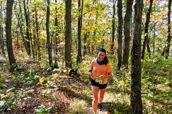 A woman wih bib number 941 wears a peach shirt as she runs through a forest that is lit up in fall color