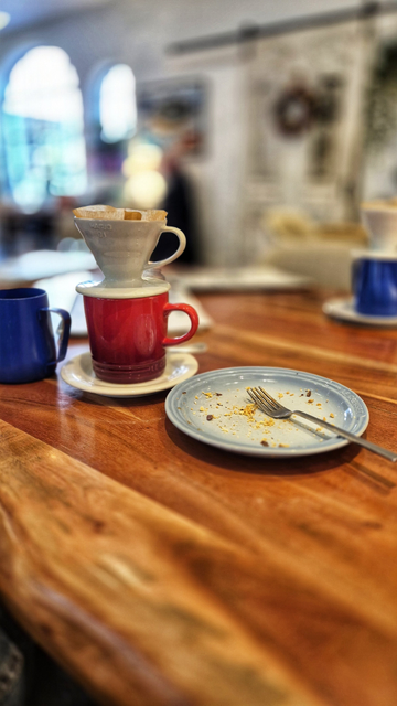 Die Szene spielt auf einer Tischplatte aus reichlich gemasertem Holz. Die Aufnahme entstand fast auf Höhe der abgebildeten Gegenstände. Auf der Tischplatte stehen ein gebrauchter Kuchenteller mit Gabel, links davon ein weißer Unterteller, darauf ein roter Kaffeebecher und darauf ein weißer Kaffeefilteraufsatz. Links davon steht ein blauer Kaffeebecher. Rechts davon, etwas entfernt und unscharf, ist ein weiterer blauer Kaffeebecher auf einem weißen Unterteller zu sehen, auf dem ein weißer Kaffeefilteraufsatz steht. Der Hintergrund ist unscharf. 