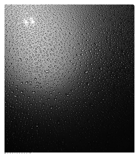 Aufnahme in schwarzweiß, tausende kleine Wassertropfen vom letzten Regen auf einer sich stark spiegelnden Motorhaube bei Nacht, ebenso spiegelt sich eine Straßenlaterne wieder  