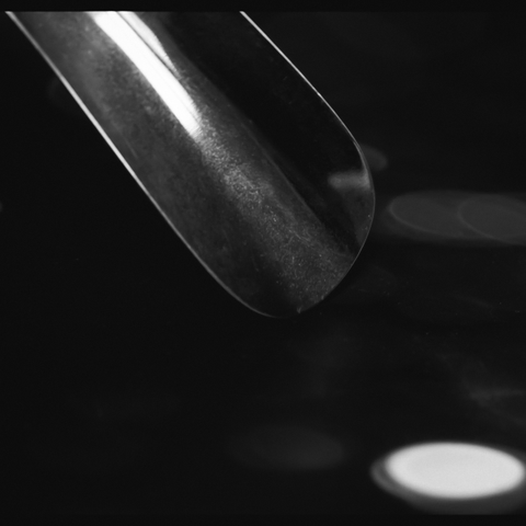 Zu sehen ist ein Schuhlöffel aus Metal, der von links oben in der Ecke des Fotos diagonal in die Bildmitte führt. An dem sonst schwarzen Hintergrund sieht man einige runde Objekte, eine davon scheint gerade erst vom Schuhlöffel runter gefallen zu sein so hell und weiß ist dieses runde Objekt. Das ganze nFoto ist in Schwarzweiß.