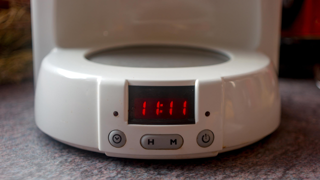Front view of a coffee machine with a clock, showing 11:11 in focus, and blurring to the back. There is no pot on the heat plate.