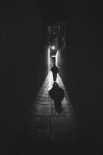 The image is a black and white photograph depicting a narrow alleyway at night. The scene is dominated by the contrasting interplay of light and shadow, creating a strong sense of mood and atmosphere.

The alley is enclosed by two brick walls that recede into the distance, converging towards a distant opening. The bricks are textured and appear to be aged, adding to the gritty realism of the scene. The ground is paved with rectangular stone slabs, some of which reflect the light, suggesting a recent rain or dampness.

A silhouetted figure is walking towards the end of the alley. The person is wearing a hooded jacket, obscuring their face and identity. Their long shadow stretches out in front of them, cast by a light source positioned behind them. The shadow adds a dramatic element to the composition, emphasizing the figure's isolation and the overall sense of mystery.