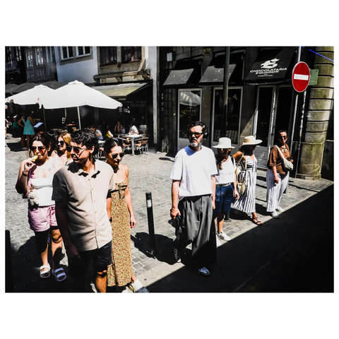The image depicts a busy urban street scene during daylight. A group of people is standing on a cobblestone sidewalk, waiting to cross the street. Most people are looking in the same direction. The individuals are dressed in casual summer attire, including shorts, dresses, hats, and sunglasses. Some people are carrying bags or holding items.

In the background, there is an outdoor seating area with tables and large white umbrellas, likely belonging to a café or restaurant. The storefronts have signs, and one of the signs reads "Chocolataria Equador," indicating a chocolate shop. The street is lined with buildings featuring traditional architectural elements. A "no entry" traffic sign is visible on the right side of the image. (draft by mistral.ai, edited by author)