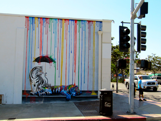 The image shows a street scene with a vibrant mural of a tiger on the side of a building. The tiger is depicted lying down with its head resting on its paws, and a black umbrella is positioned above it. The building's exterior is bright white, and the mural appears to have dripping paint effects in various colors like blue, green, and yellow. To the right of the mural, there's a black trash can with "Village" printed on it. In the background, a person wearing a blue shirt and khaki pants is walking, and a silver pickup truck is parked across the street, with a traffic light visible above them.