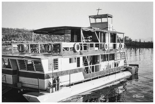 A black and white image of a vintage houseboat, docked on calm water. Life rings are visible on the side. Trees in the background.